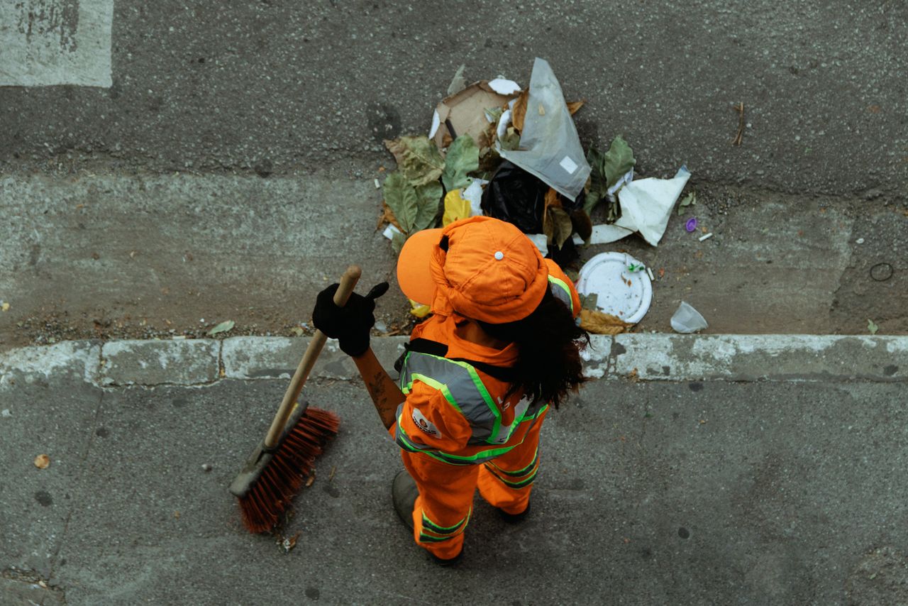 Top view of a street cleaner wearing orange uniform sweeping debris on a city sidewalk.